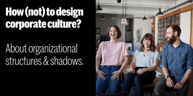 Drei lachende Personen sitzen auf einem Holztisch in einem offenen Loft-Büro; daneben der Text „About organizational structures & shadows“