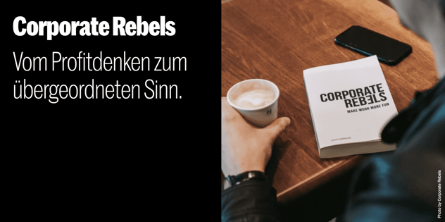 Buch „Corporate Rebels – Make Work More Fun“ auf einem Holztisch, daneben ein Smartphone und eine Hand am Kaffeebecher