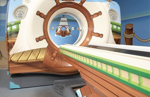 MRT-Gerät für Kinder, als Piratenschiff gestaltet: Steuerrad um die Röhrenöffnung, Holzplanken, Bullaugen und ein gemaltes Segelschiff im Inneren