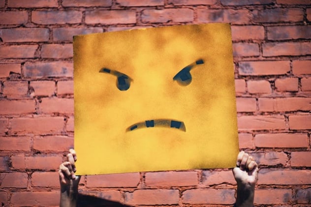 Zwei Hände halten vor einer Backsteinwand ein gelbes Pappschild mit aufgesprühtem wütendem Gesicht hoch