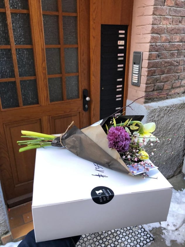 Weiße Frühstücksbox mit Aufkleber „Bock auf Frühstück“ und aufgelegtem Blumenstrauß, abgestellt vor einer Altbau-Haustür