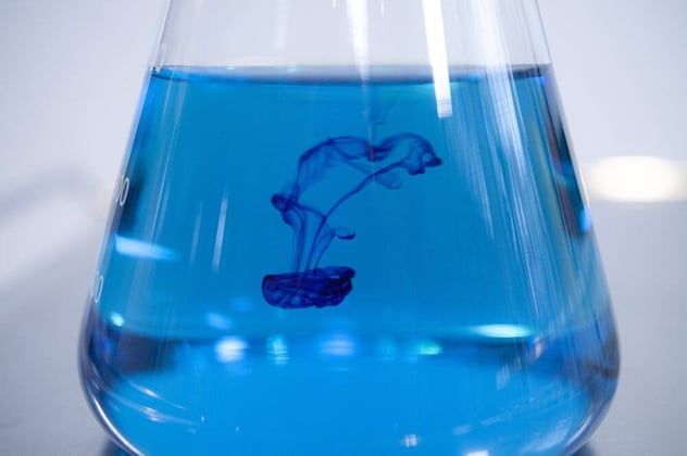 Dunkelblaue Farbtropfen verteilen sich schlierenförmig in der blauen Flüssigkeit eines Erlenmeyerkolbens