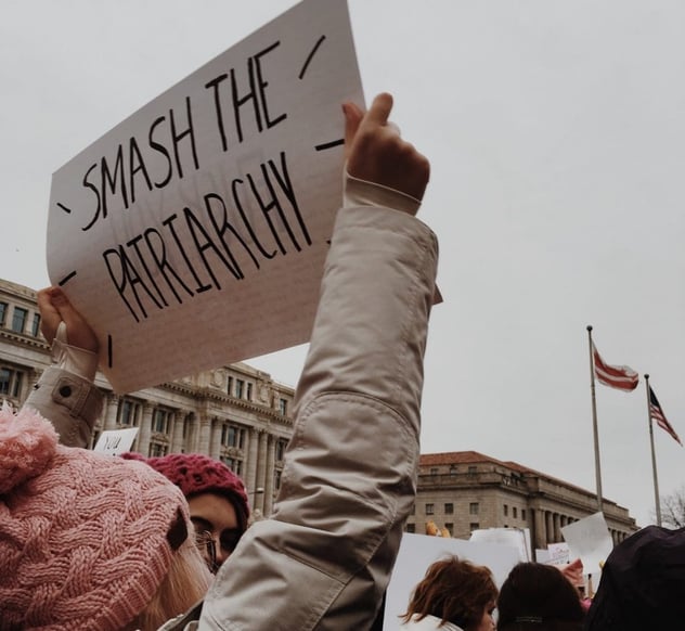 Demonstrantin hält auf einer Kundgebung ein handgeschriebenes Pappschild mit der Aufschrift „Smash the Patriarchy" hoch, im Hintergrund pinke Mützen und weitere Schilder