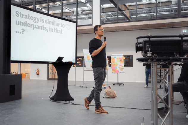Redner mit Mikrofon auf einer Bühne vor der Präsentationsfolie „Strategy is similar to underpants, in that …?" mit Unterhosen-Icon; im Hintergrund Flipcharts voller Klebezettel