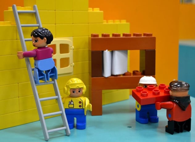 Vier Lego-Figuren bauen gemeinsam an einer gelben Mauer: eine steigt die Leiter hoch, eine trägt Bausteine heran, zwei stimmen sich am Gerüst ab