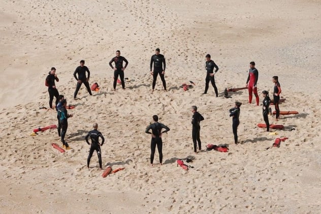 Gruppe von Rettungsschwimmer*innen in Neoprenanzügen steht im Kreis am Strand, Rettungsbojen und Ausrüstung liegen im Sand