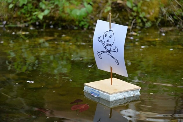 Selbstgebautes Spielzeugboot aus Styropor, Holzplatte und Papiersegel mit gemaltem Totenkopf treibt auf einem Teich