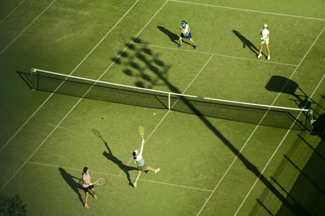 Vier Tennisspielerinnen und -spieler im Doppel auf einem Rasenplatz, aus der Vogelperspektive im Moment des Aufschlags